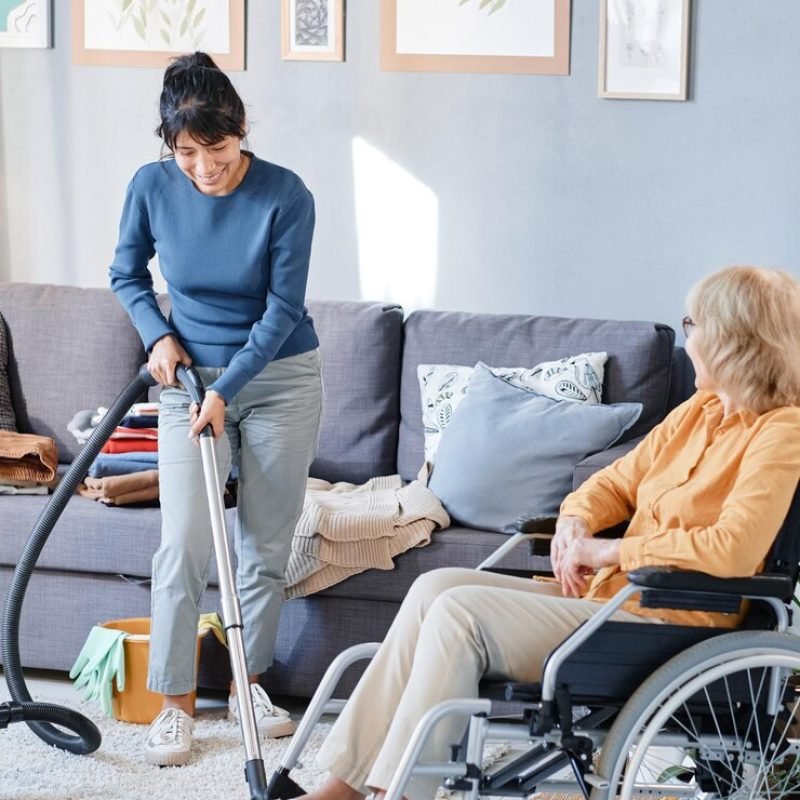 senior-woman-using-wheelchair-talking-housekeeper-who-vacuuming-floor-room_249974-10828